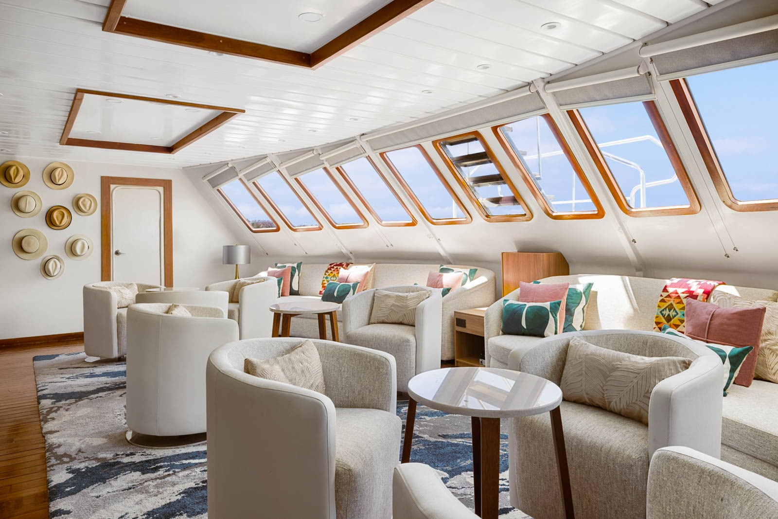 Cozy lounge with white chairs, colorful pillows, and hats on the wall, sunlight streaming through angled windows.