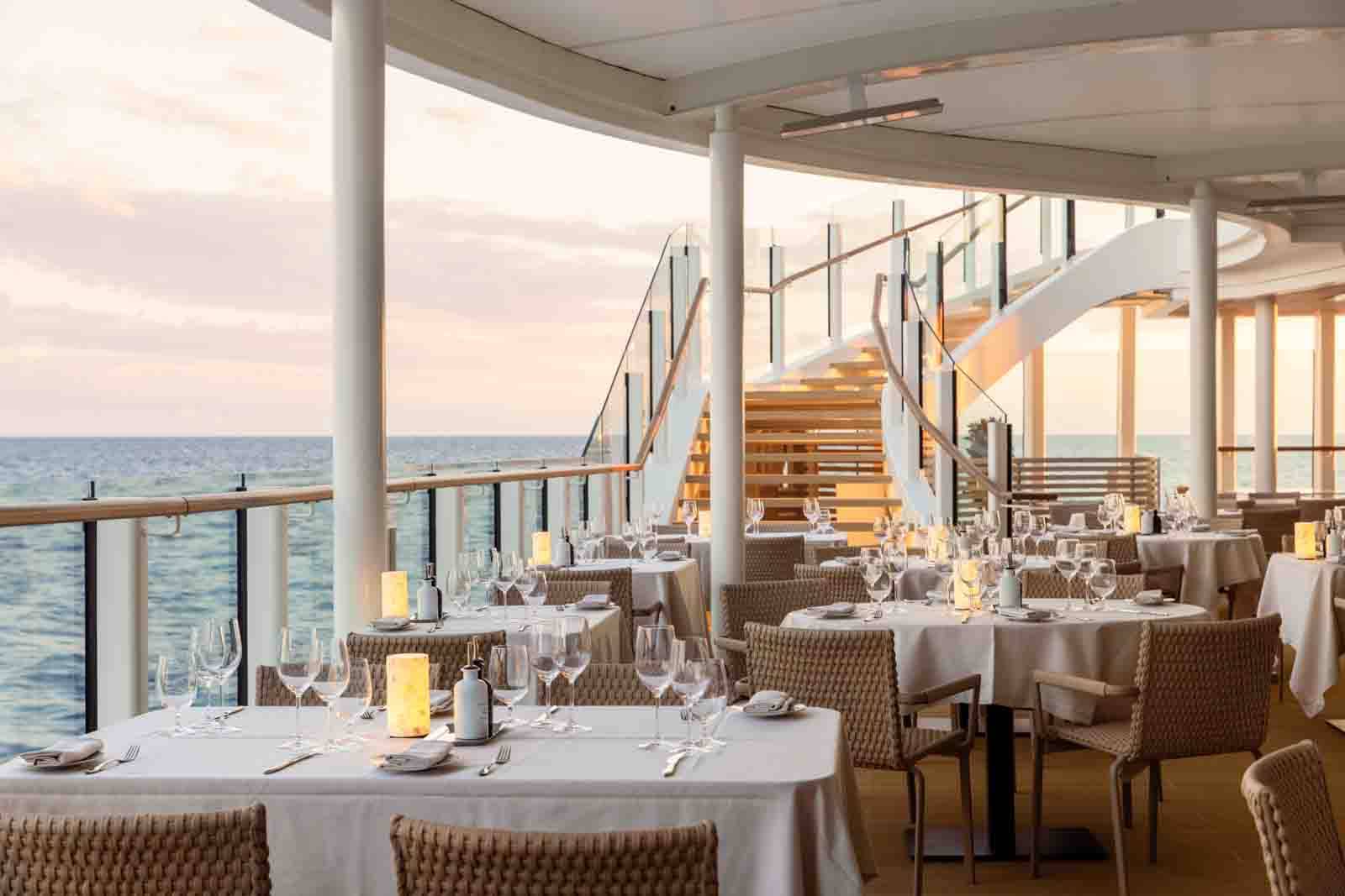 Elegant outdoor dining area with white tablecloths, tan chairs, and glowing lamps, beside the ocean at sunset.