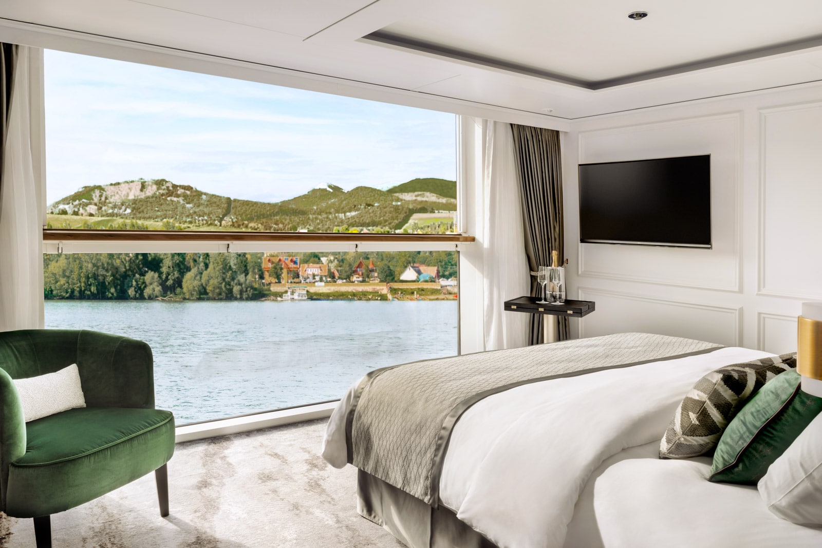 Modern cruise suite with green chair, white bed, and large window showing blue water and green hills under a pale sky.