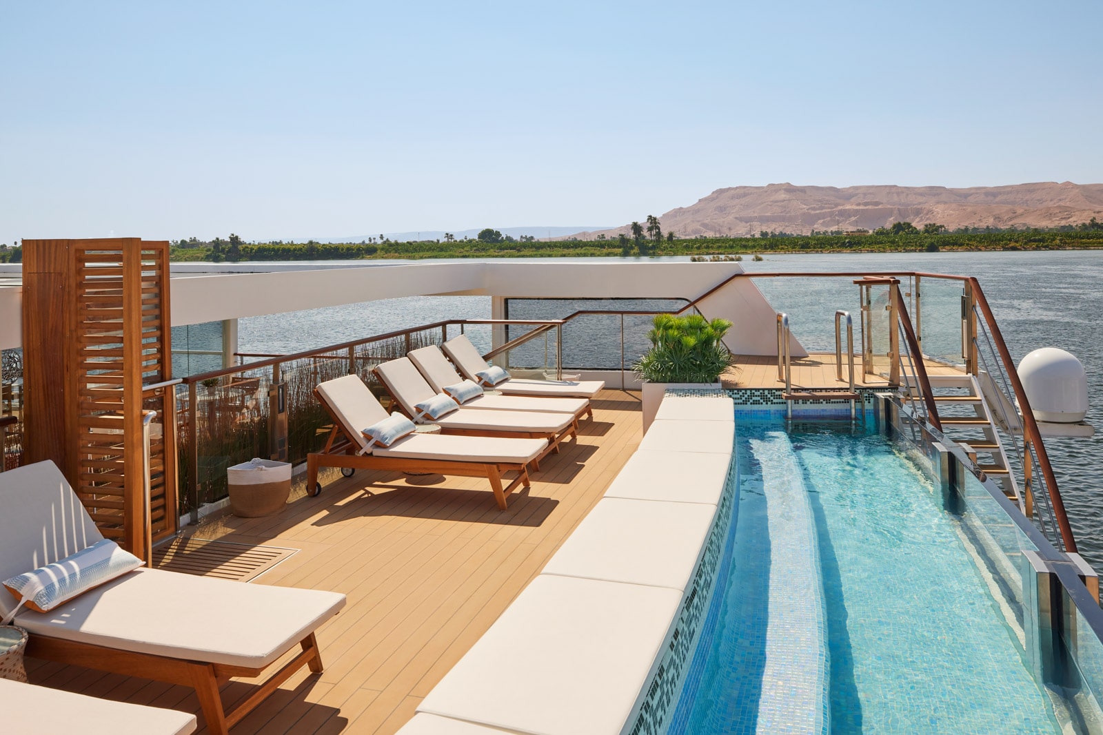 Sunlit pool deck with blue water, tan loungers, and wood accents, overlooking a river and sandy hills in the distance.