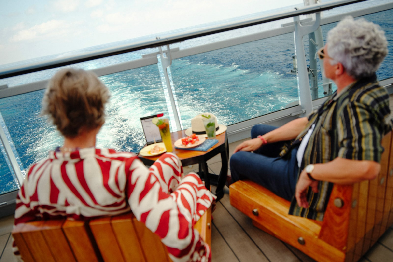 Two people sit on deck chairs staring out at the sea from a Virgin Voyages cruise ship.