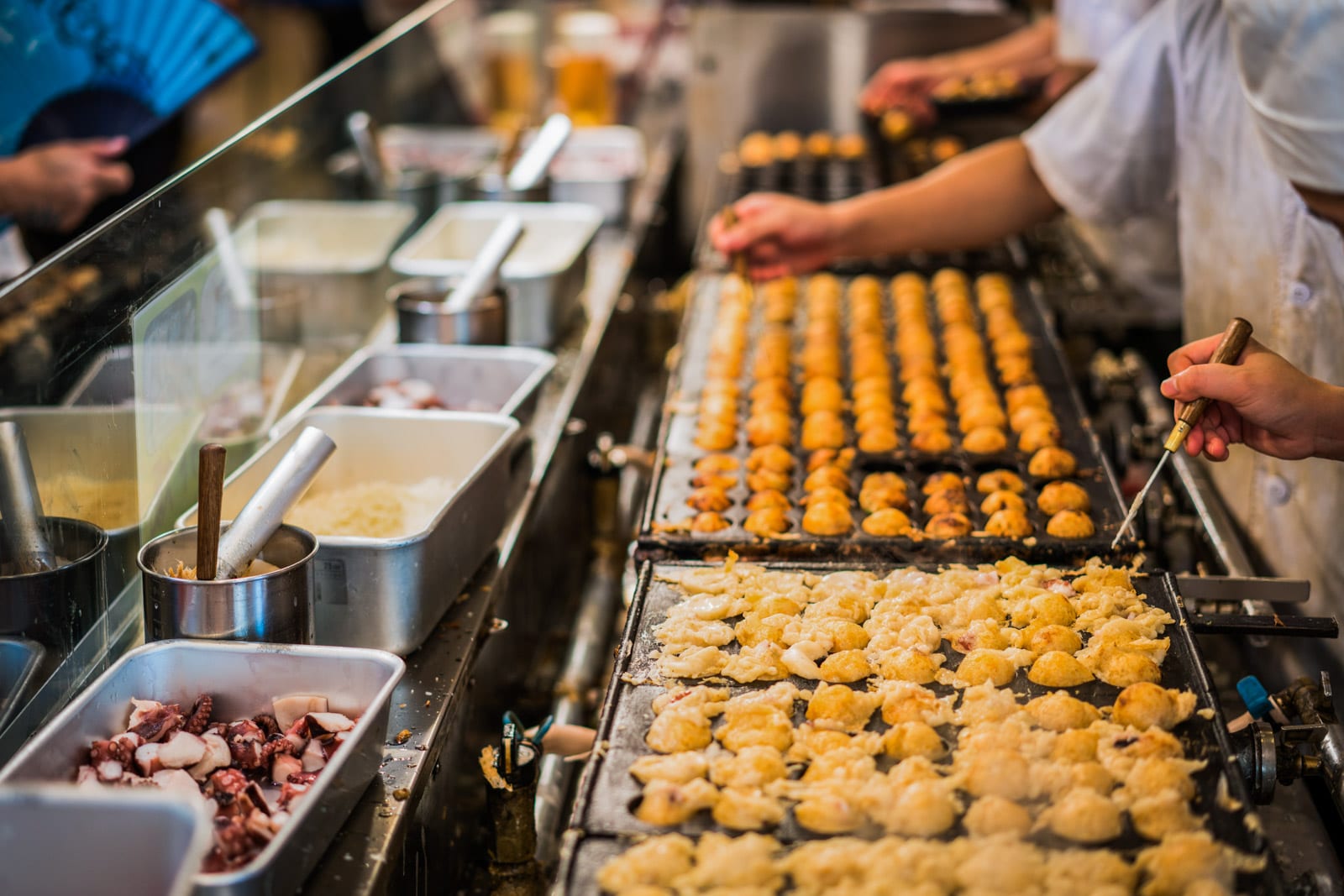 Chefs preparing takoyaki balls on a grill with trays of ingredients in a busy kitchen.