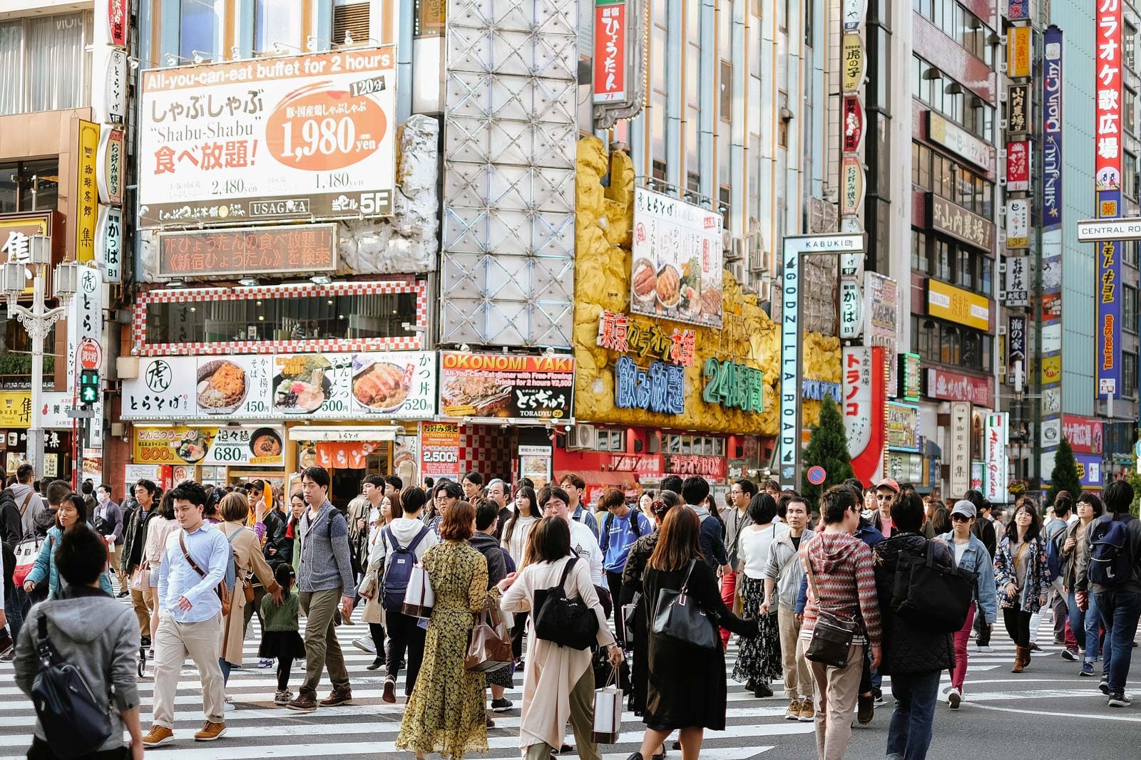 People walking across a city street lined with colorful signs and restaurant advertisements.