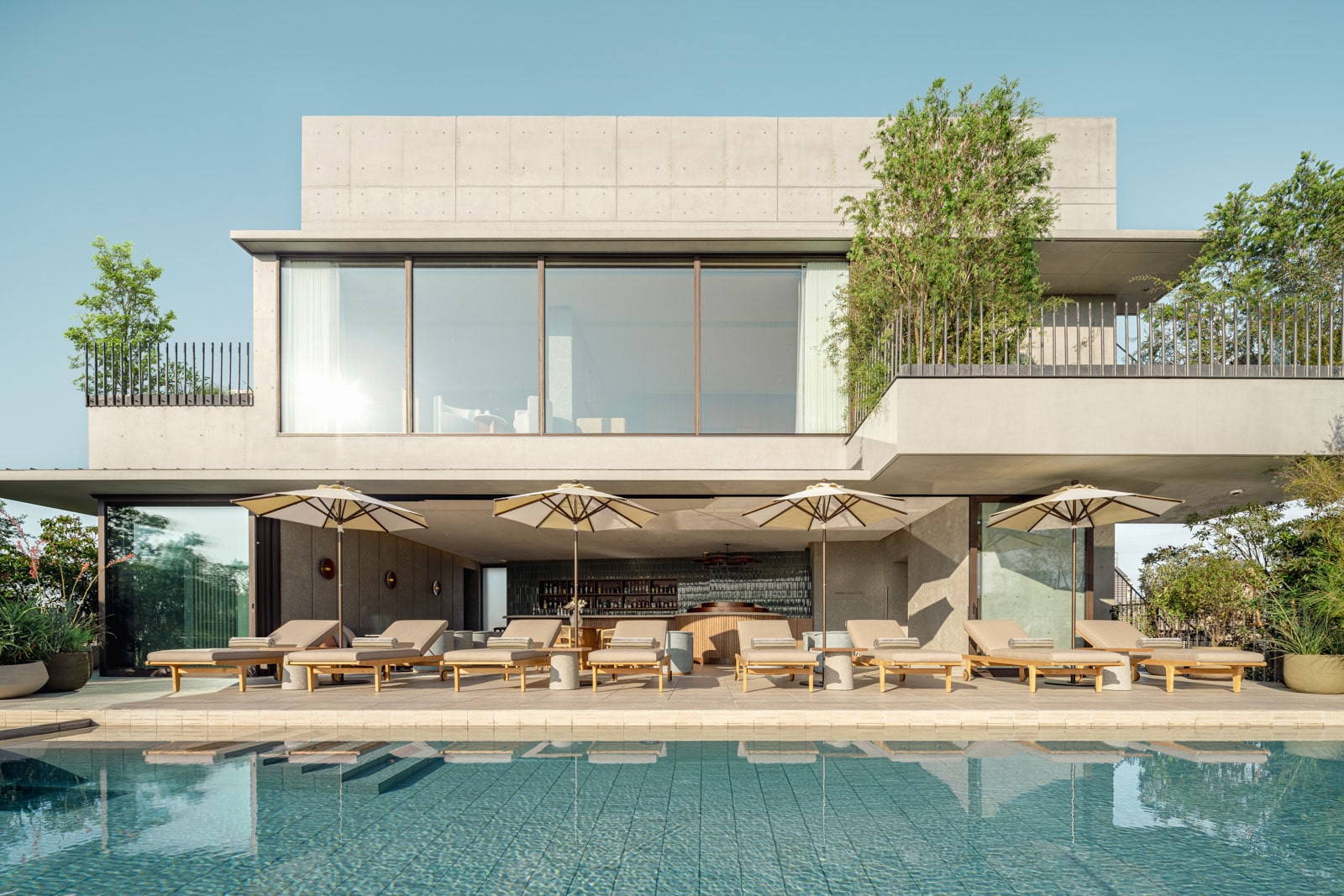 Modern villa with poolside lounge chairs, umbrellas, and large windows surrounded by greenery.
